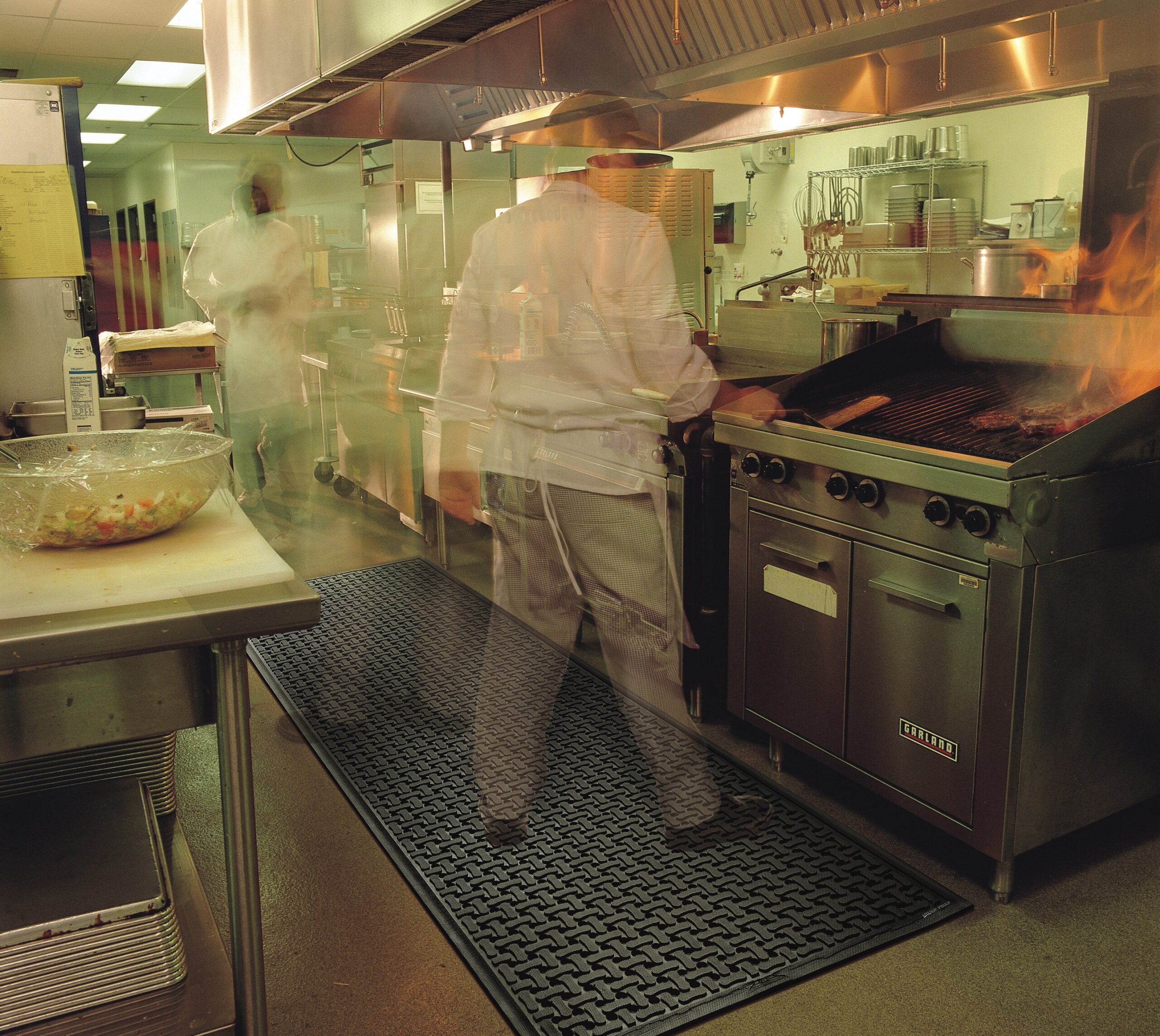 Chefs working in a busy commercial kitchen with flames on the grill, stainless steel equipment, and anti-slip floor mats to ensure safety despite the fast-paced environment.