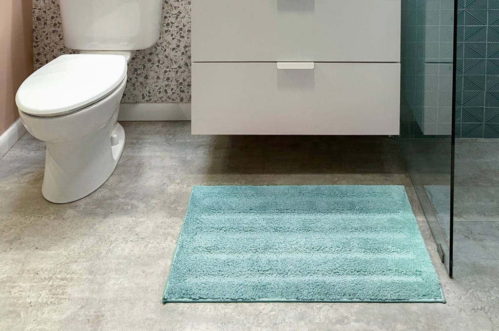 Modern bathroom with a clean design, featuring a white toilet, vanity, and a soft teal bath mat on a tiled floor.