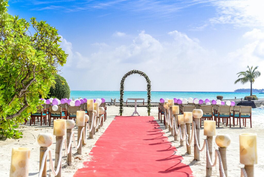 Beachfront wedding setup with a red carpet aisle, candle-lined path, and an arch overlooking the ocean under a clear blue sky.