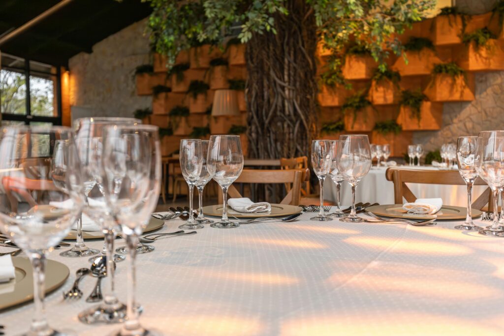 Outdoor dining setup with a round table covered in a clean white tablecloth, surrounded by wooden chairs, glassware, and a tree with decorative shelves in the background.