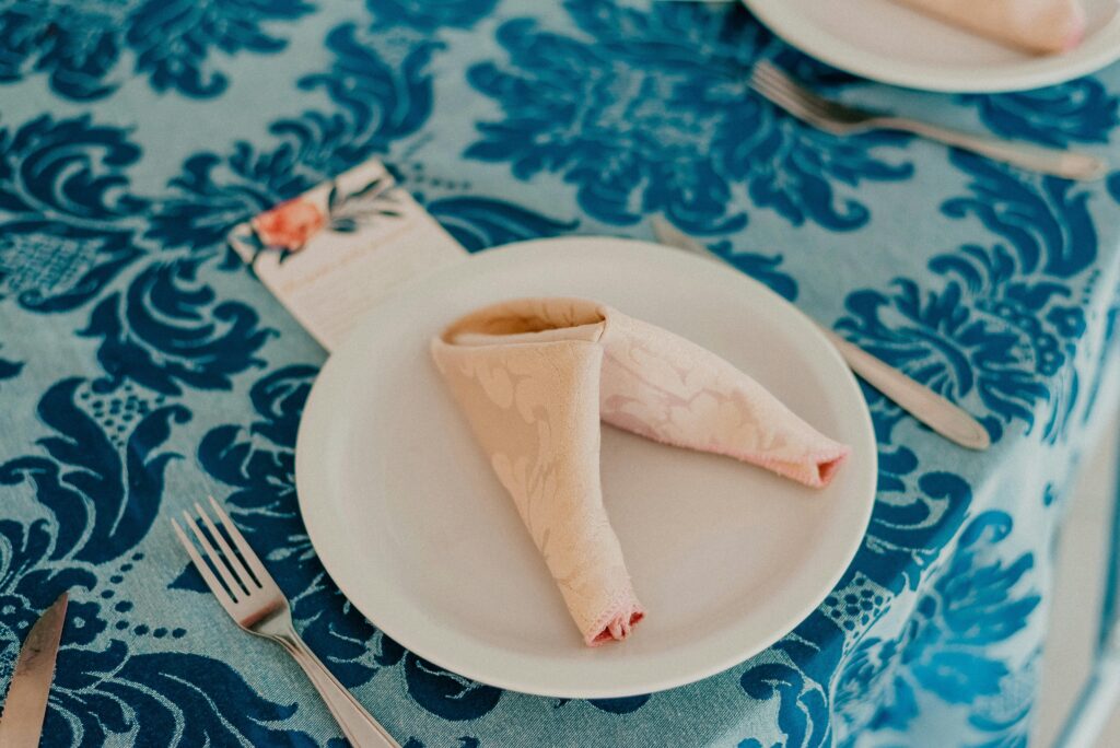 Close-up of a table setting with a folded napkin on a white plate, placed on a blue patterned linen tablecloth.