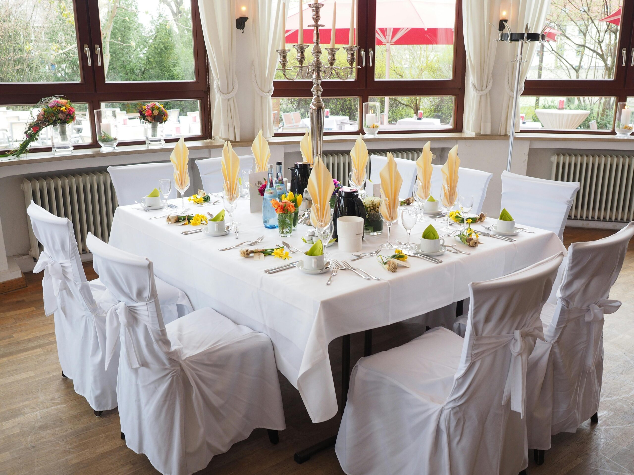 Indoor dining table with white linens, chair covers, and neatly folded napkins, accented by floral centerpieces and natural light.