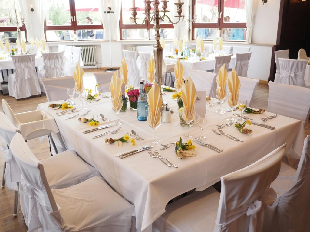 Elegant dining setup with white tablecloths, yellow napkins, floral centerpieces, and chairs covered in white covers with sashes, in a bright room with large windows.