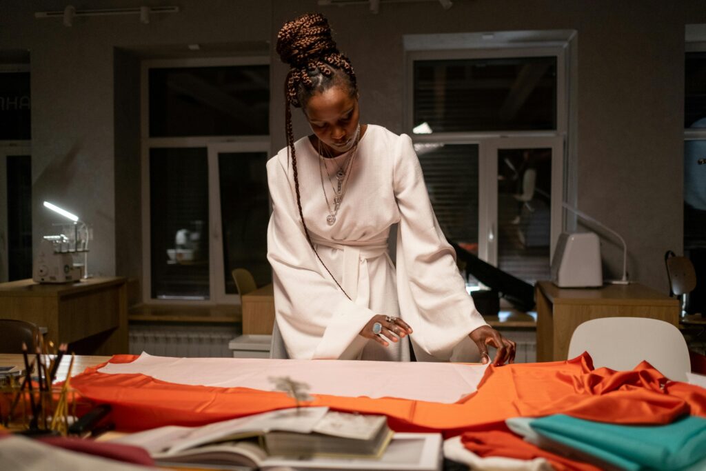 Lady in a white outfit working on fabric patterns at a table, surrounded by colorful fabrics and abilities in a studio at night.