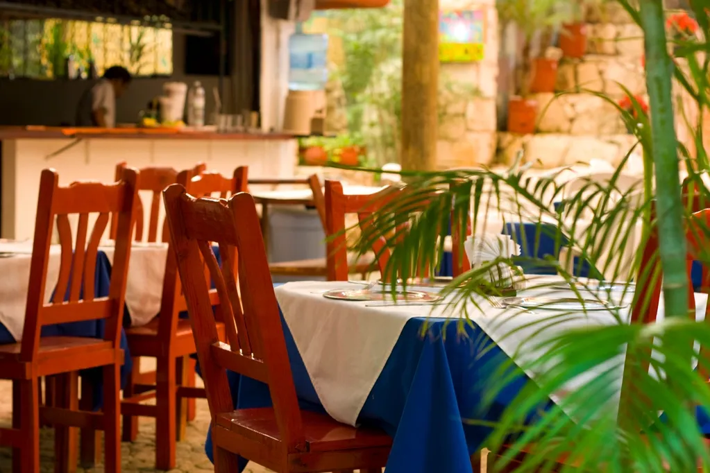 Outdoor restaurant with wooden chairs and tables covered in white and blue tablecloths, surrounded by greenery.