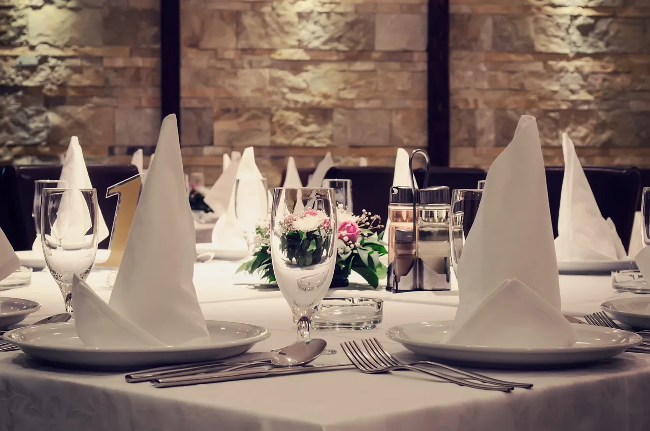 Elegant table setting with white napkins folded into triangles, plates, glasses, cutlery, and a floral centerpiece, in a dining area with a stone wall background.