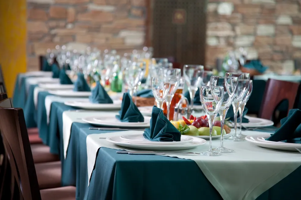 Elegant dining setup with long tables covered in white and green tablecloths, decorated with wine glasses, napkins, and centerpieces.