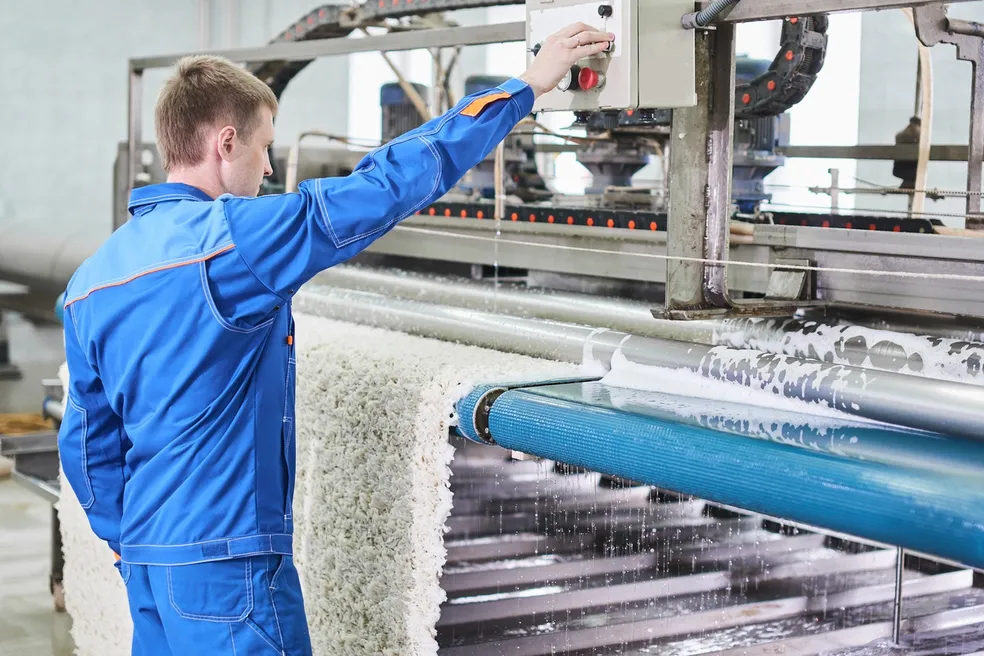 Worker in a blue uniform operating an industrial machine processing fabric or textiles in a manufacturing facility.